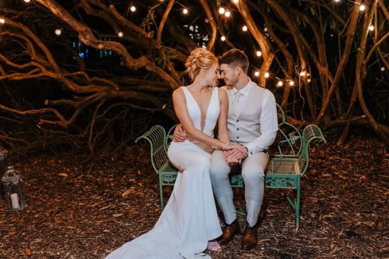 Elegant bride and groom sitting close on a garden bench at night with string lights, celebrating love and marriage.