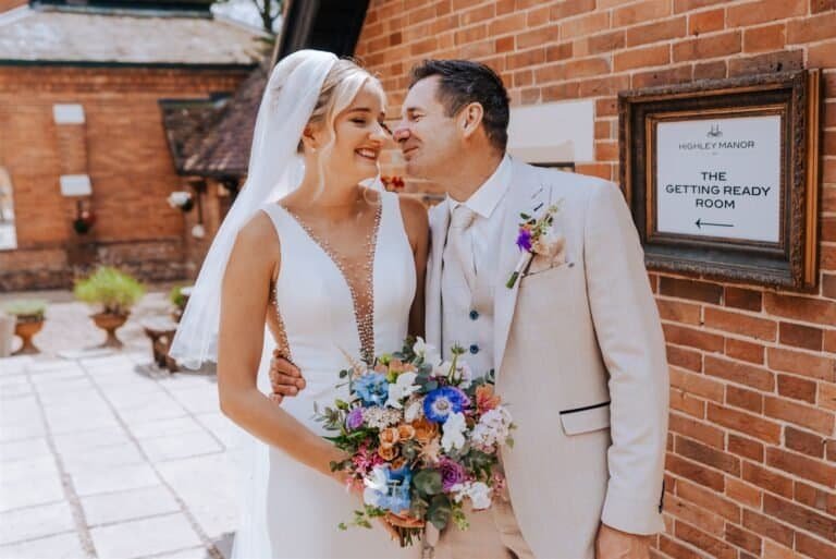 Bride and groom smiling at each other outside the brick barn, the bride holding a bright summer bouquet