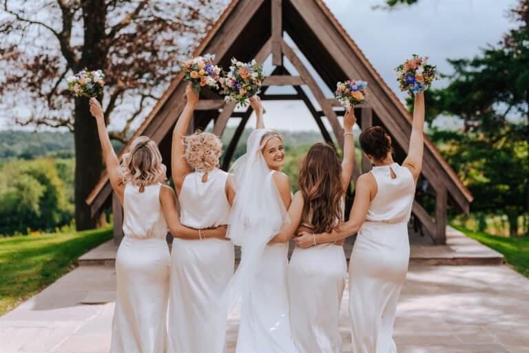 Beautiful bride with bridesmaids holding bouquets outdoors at a rustic wedding venue.