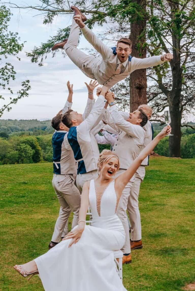 Vibrant wedding celebration outdoors with a bridal party lifting a groom, surrounded by lush greenery.